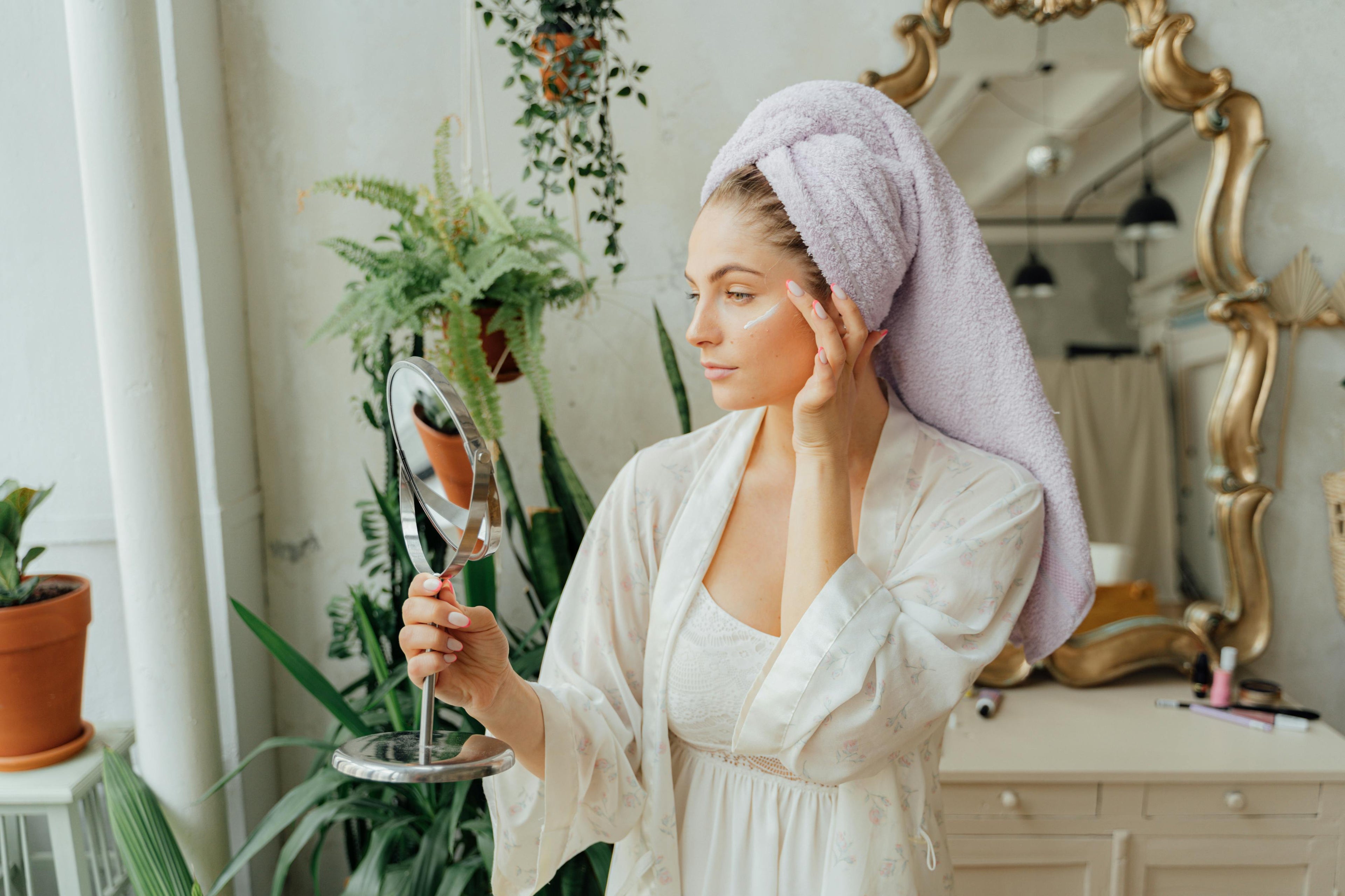 Woman in a white robe with a towel on her head, holding a mirror in a decorated room.